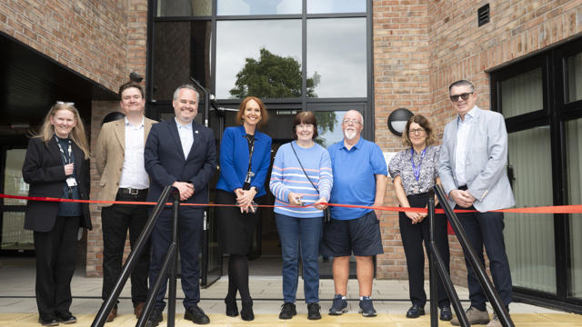 Collection of people in front of building with a red ribbon about to be cut