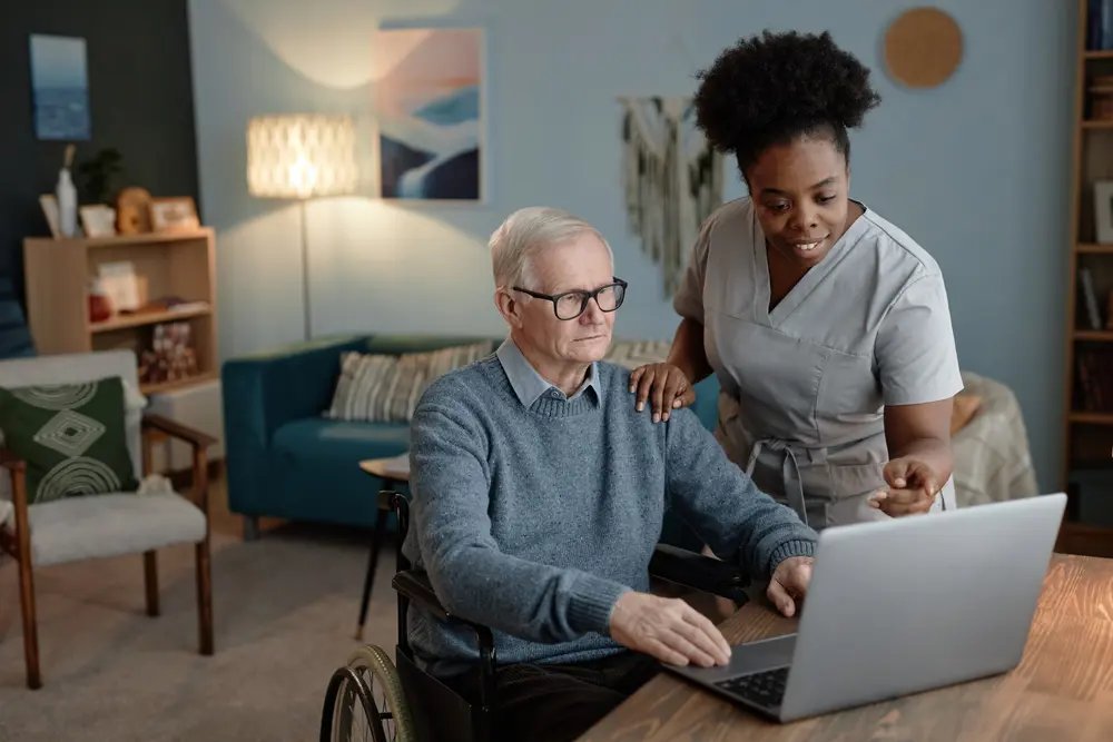 Older man in a wheelchair getting help using a laptop by a woman 