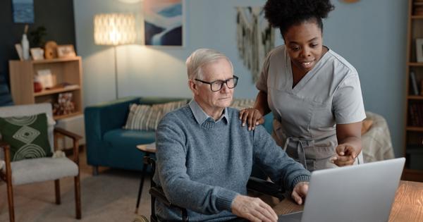 Older man in a wheelchair getting help using a laptop by a woman 