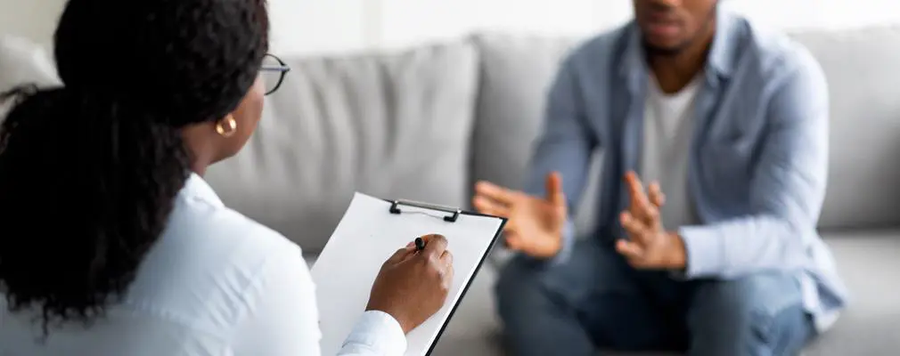 A woman holding a notepad speaking to a man on the sofa