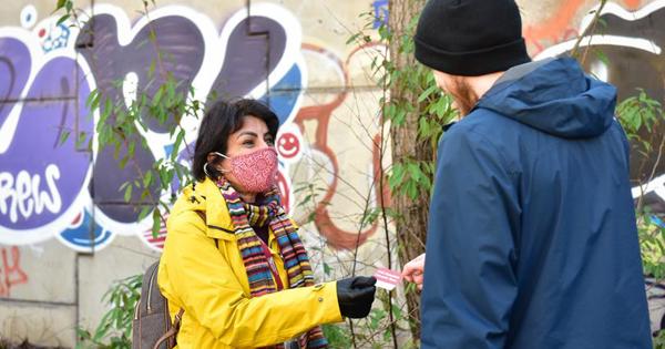 A woman in a yellow jacket and face mask handing out a card to a man on the street
